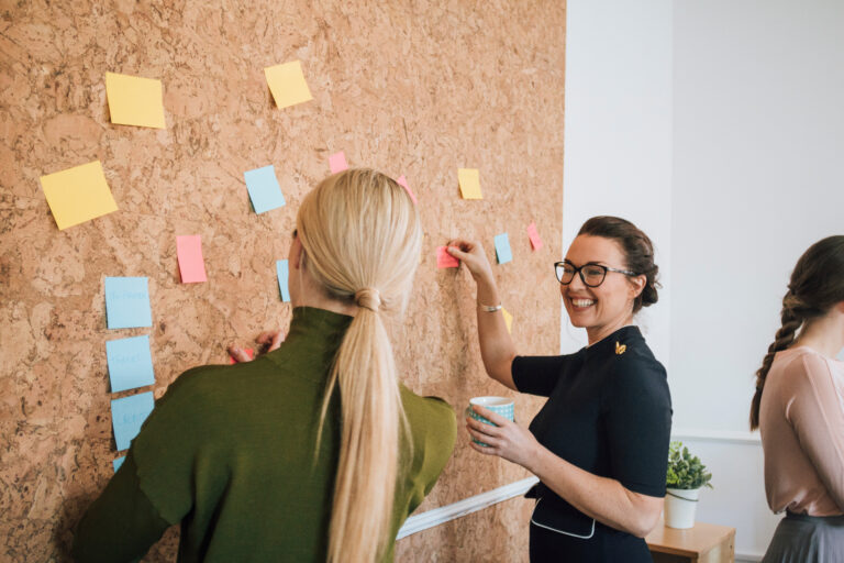 Two women are at work in an office. They are standing at a cork board and are having a discussion as they pin things up, Getting more done in less time