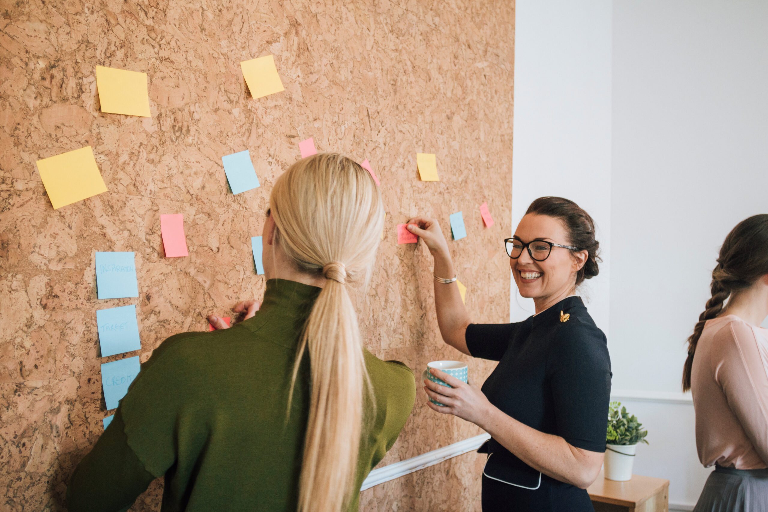 Two women are at work in an office. They are standing at a cork board and are having a discussion as they pin things up, Getting more done in less time