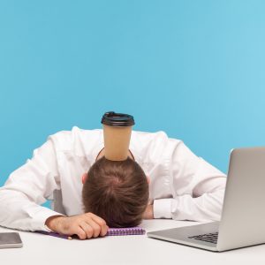 Overworked exhausted man office worker lying on table, paper coffee cup standing on his head, lack of energy, procrastination and professional burnout