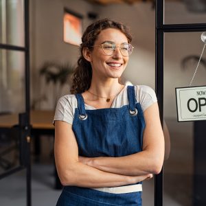 Smiling small business owner showing open sign in her shop while leaning against the door, startup business