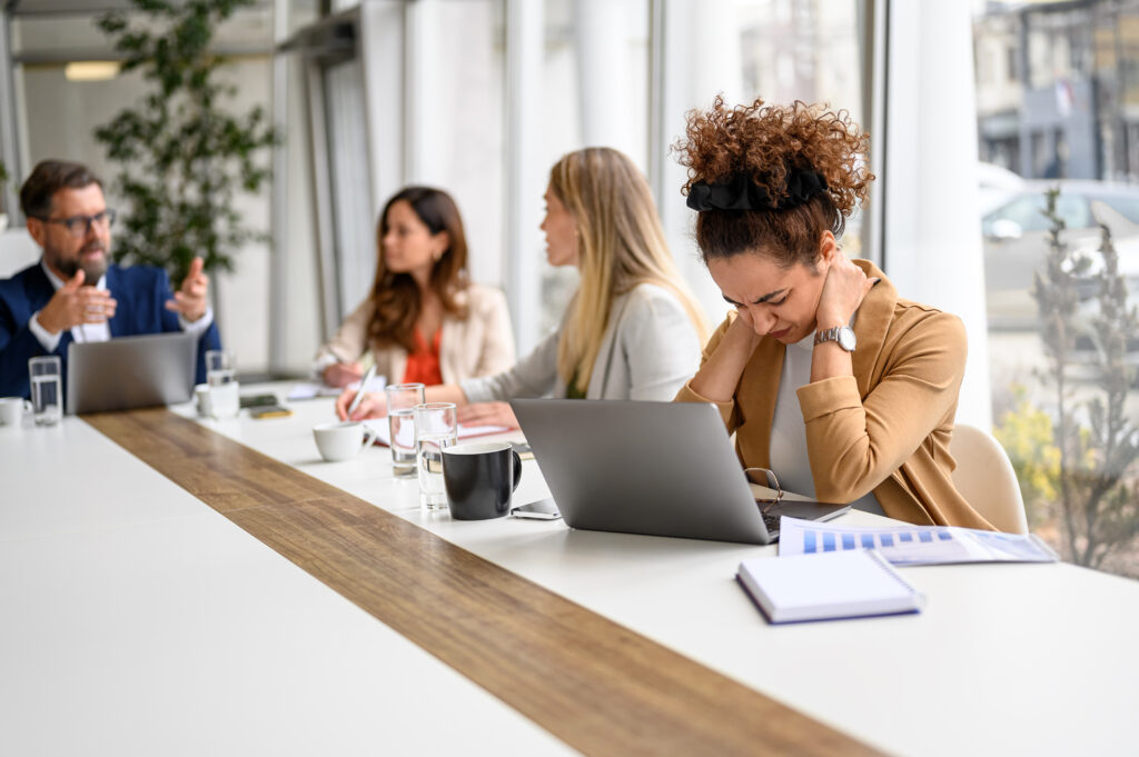 Stressed businesswoman holding her neck in pain and working on laptop while colleagues discussing in meeting, having new tasks, Time