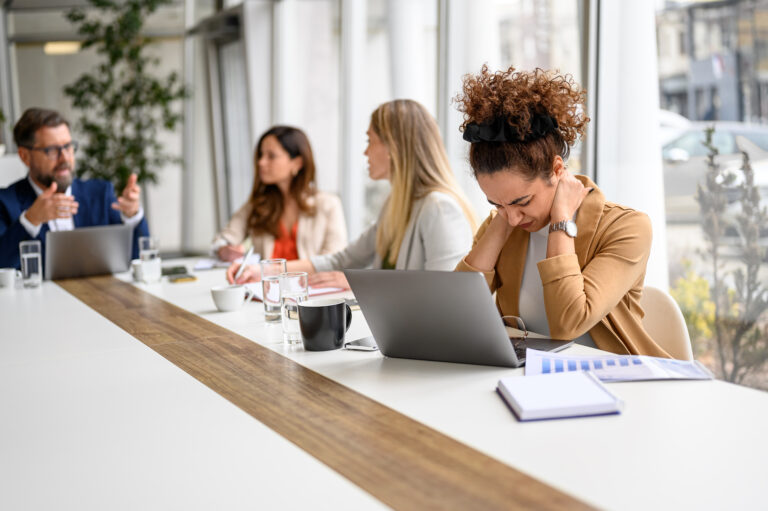 Stressed businesswoman holding her neck in pain and working on laptop while colleagues discussing in meeting, having new tasks, Time