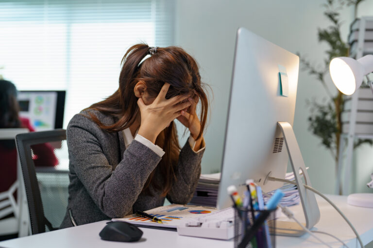 Young professional woman experiencing stress and burnout while working on a computer in a modern office, covering her face with hands and struggling with a difficult task or deadline