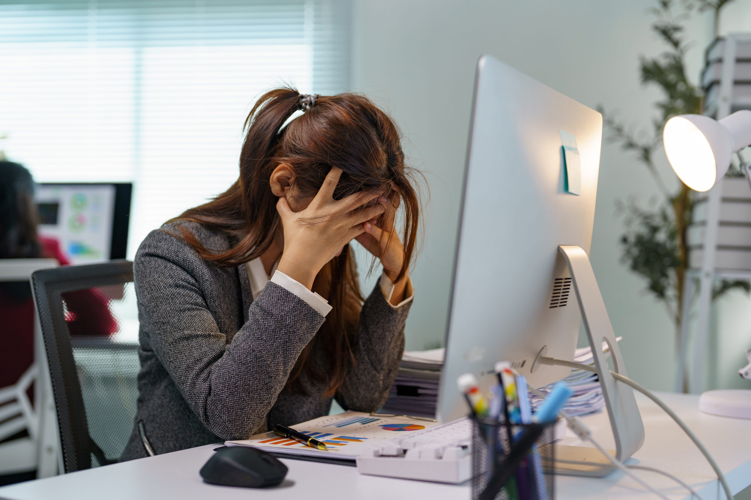 Young professional woman experiencing stress and burnout while working on a computer in a modern office, covering her face with hands and struggling with a difficult task or deadline