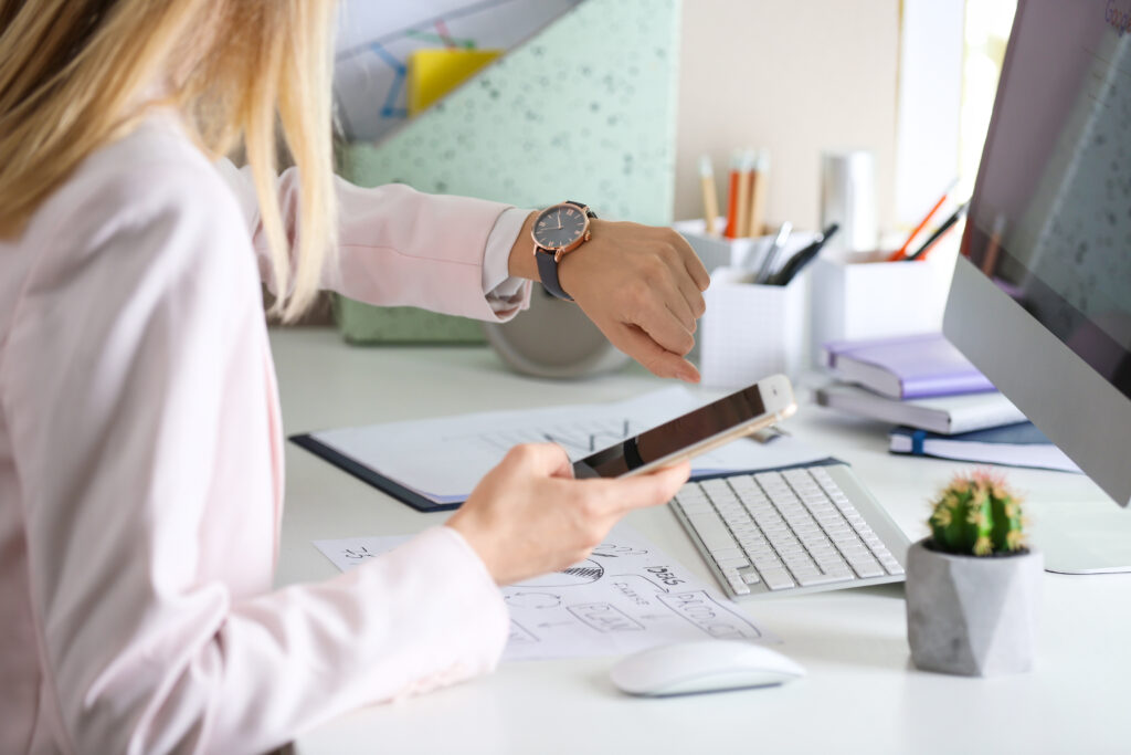 Young woman checking time on her wristwatch at workplace, wasting hours by doing these things at work