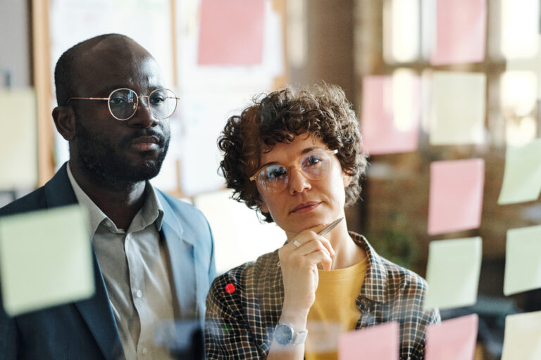 Two employees in eyeglasses reading plans together on sticky notes hanging on glass wall at office, the Art of Accountability