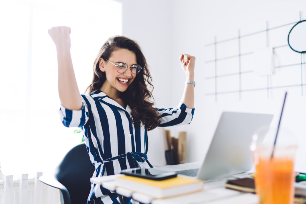 Overjoyed young woman sitting at desk with laptop with raised arms in winner gesture and celebrating achievements, win more in your business