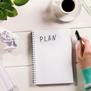 Notebook on office table with a cup of coffee, plant, note and paper. woman's hand wrote the word PLAN. Top view desk, flat lay, plan writing top view