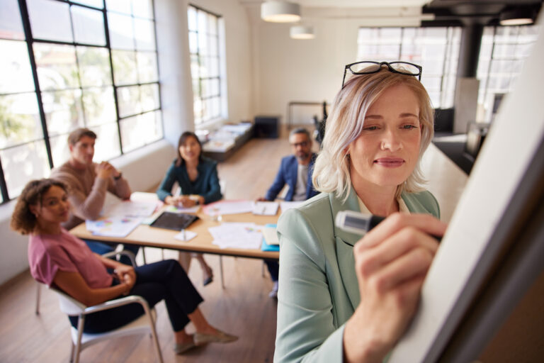 Mid adult woman leading a team meeting, Team members watching a presentation by their boss on a whiteboard. Mature woman writing strategy while sharing marketing goals and planning with collaborative business team sitting in background, Ways to Coach Your Employees to Greatness