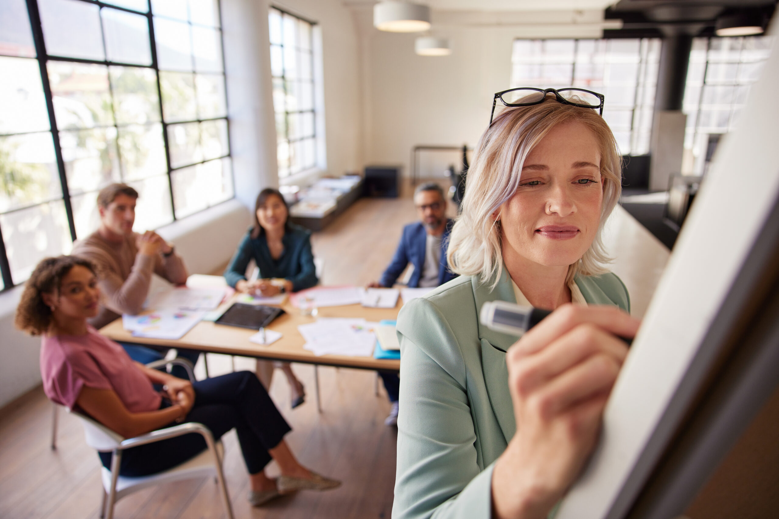 Mid adult woman leading a team meeting, Team members watching a presentation by their boss on a whiteboard. Mature woman writing strategy while sharing marketing goals and planning with collaborative business team sitting in background, Ways to Coach Your Employees to Greatness