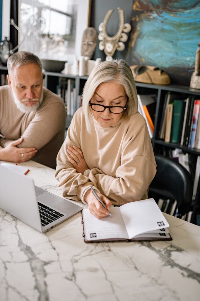 Elderly couple using a laptop and writing in a notebook in a cozy home setting.